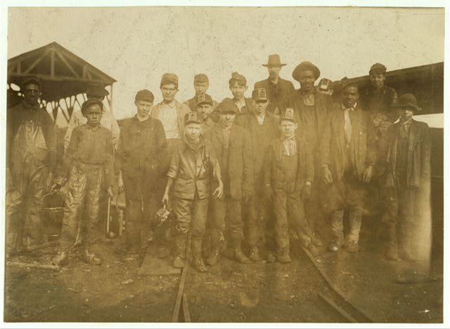 About eighteen mine workers posed on the rails at the pit mouth, several of them boys, some wearing lamps on their caps, all in work clothes darkened with coal.