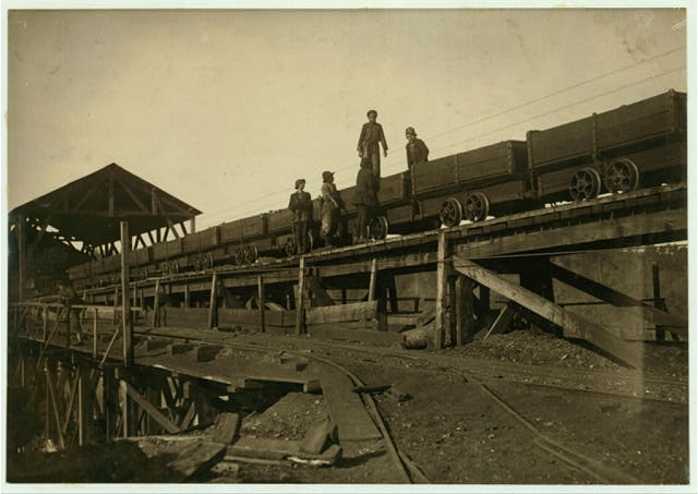 Four figures standing on an elevated wooden trestle beside a line of open coal cars, with a timber shed and railway track on the ground below.