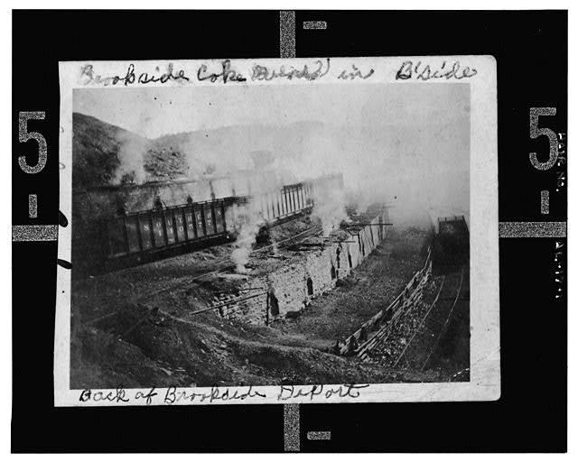 A long bank of brick coke ovens built into a hillside with smoke rising from several of them, and a line of open rail cars standing on an elevated trestle above. Handwriting on the print reads Brookside Coke Ovens in Bside, and Back of Brookside Depot.
