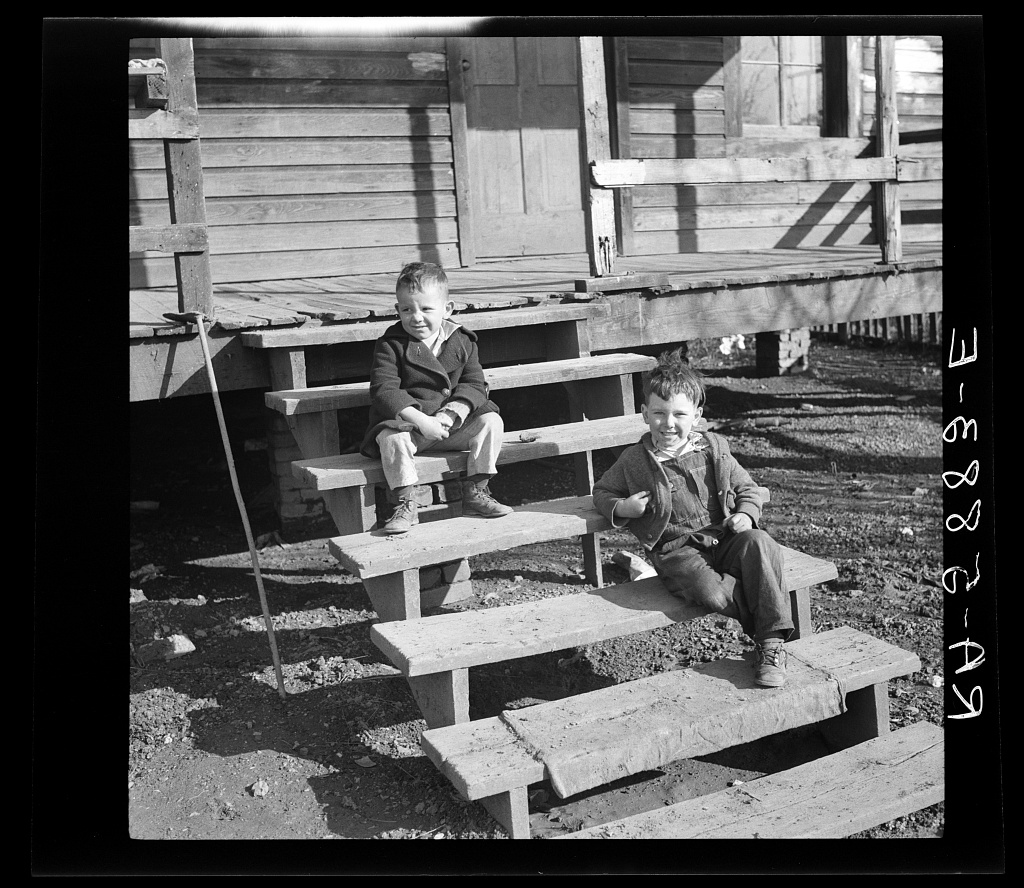 Two young boys in wool coats and work trousers sitting on the plank steps of a frame house raised on posts, both looking at the camera, with bare dirt and stones around the steps.