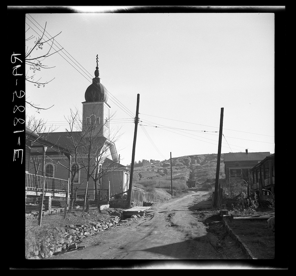 A dirt street running uphill between utility poles, with a clapboard church on the left carrying an onion dome and a three bar Orthodox cross on its tower, bare winter trees, a plank fence, frame houses on the right, and a bare eroded hillside across the valley behind.